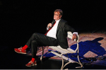 Image of Garrison Keillor reclining on a bench onstage holding a microphone while sharing a story.