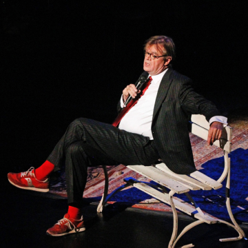 Garrison Keillor reclining on a bench onstage with a microphone