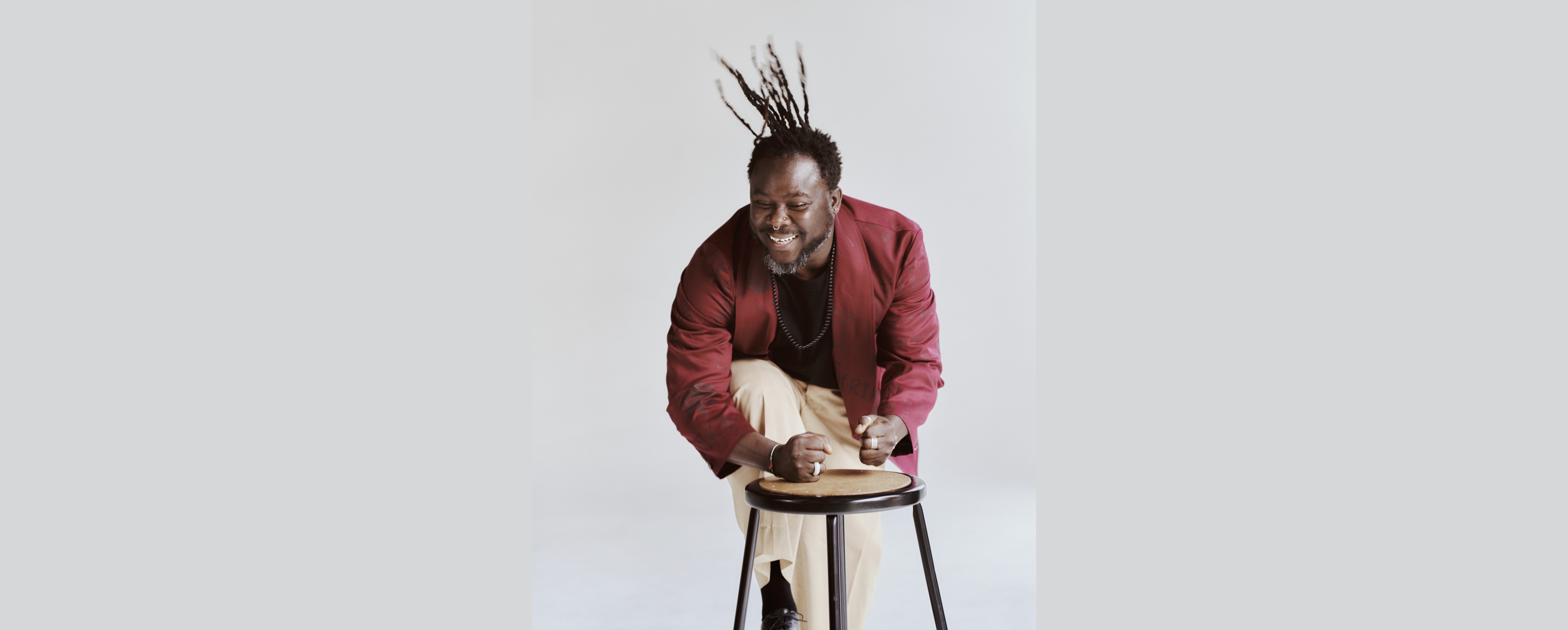 Image of Okaidja Afroso drumming on a stool with his hands, he has joyfully tossed his hair into the air