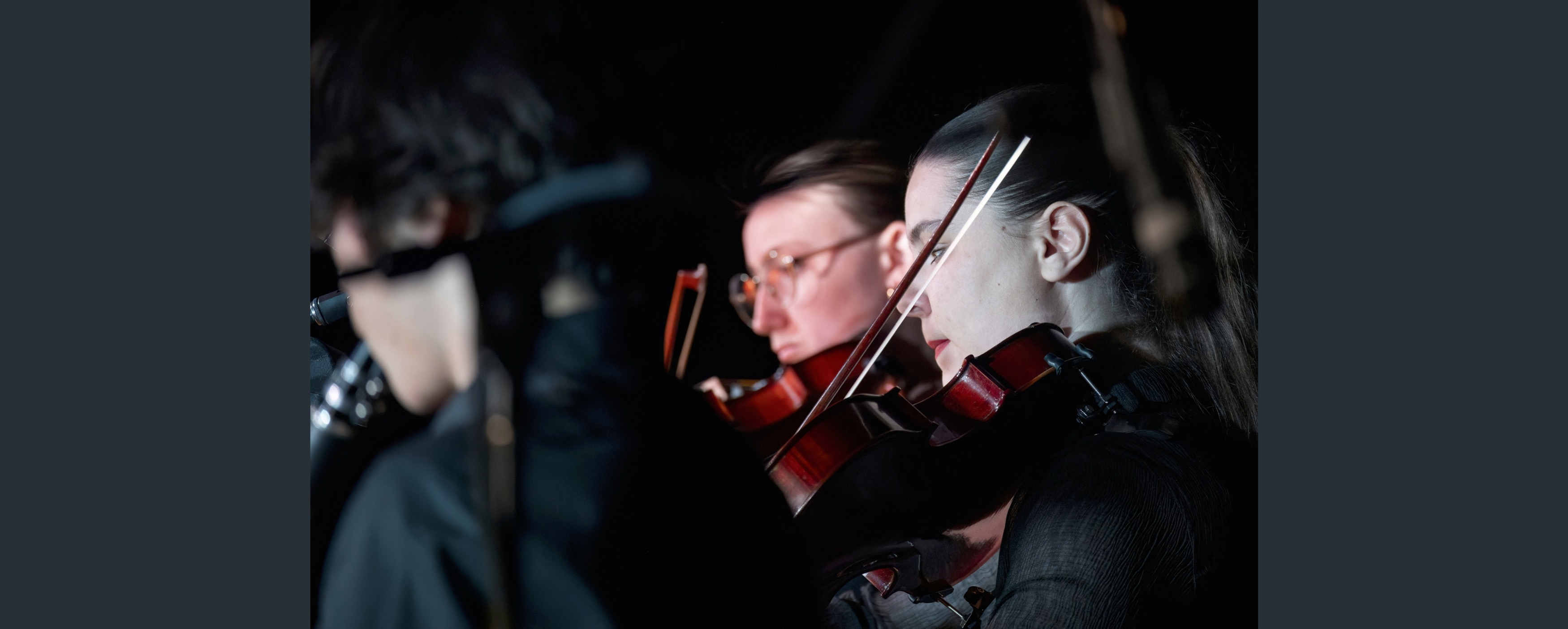 Image of three musicians. Two are out of focus and one is in focus and playing the violin with a pensive expression.