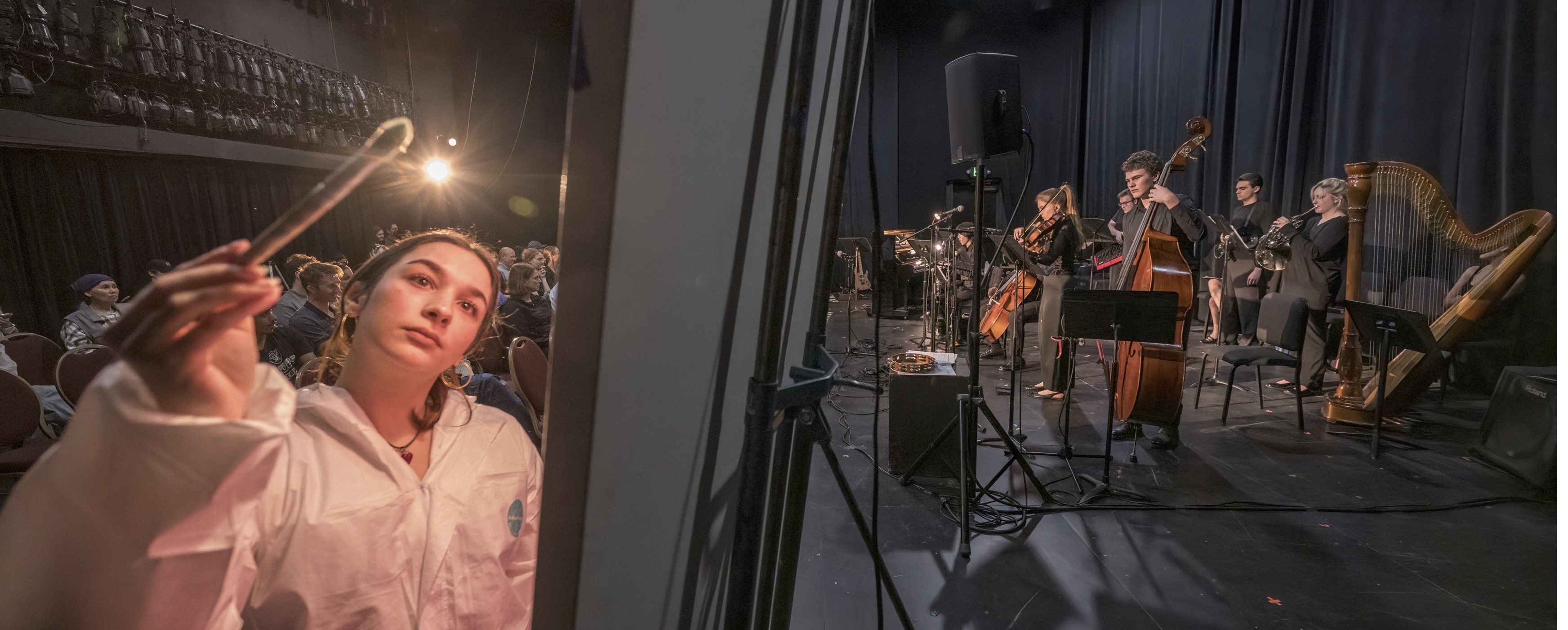 image of artist painting in the foreground with a musical ensemble in the background, all inside a blackbox theatre