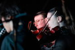 Image of three musicians, two of which are out of focus. One musician is in focus and playing the violin with a pensive expression.