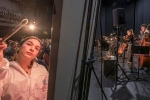 image of artist painting in foreground while musicians play onstage in the background in a blackbox theatre